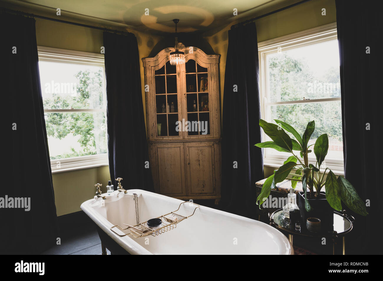 Interior view of bathroom with wooden corner cabinet  between sash windows, roll top bath with brass bath caddy. Stock Photo