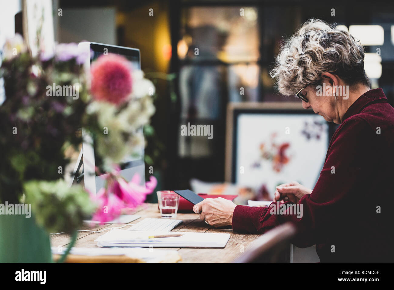 Senior woman wearing glasses and red dress sitting at a wooden table, looking at digital tablet. Stock Photo