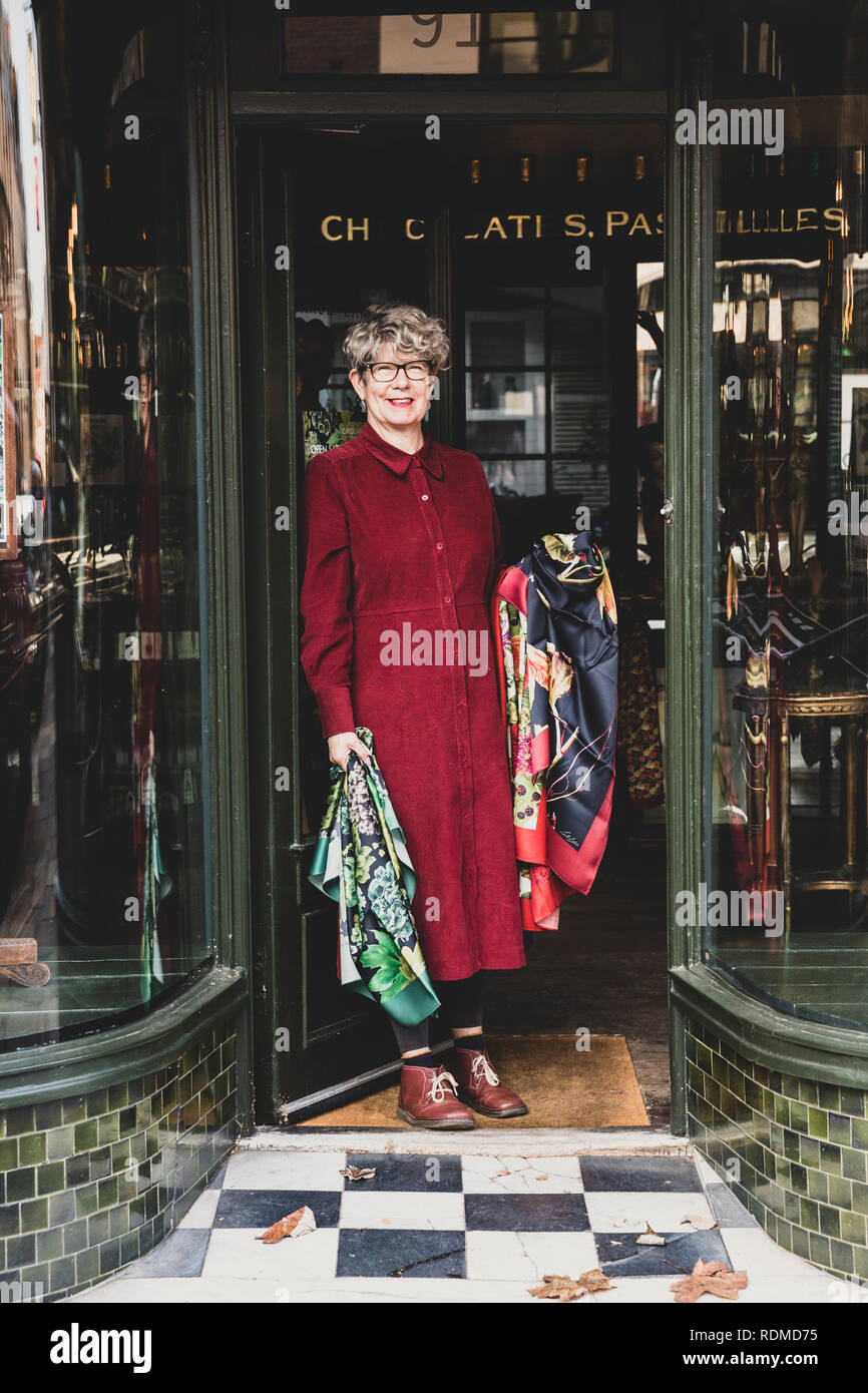 Smiling senior woman wearing glasses and red dress standing front of interior design store, looking at camera. Stock Photo