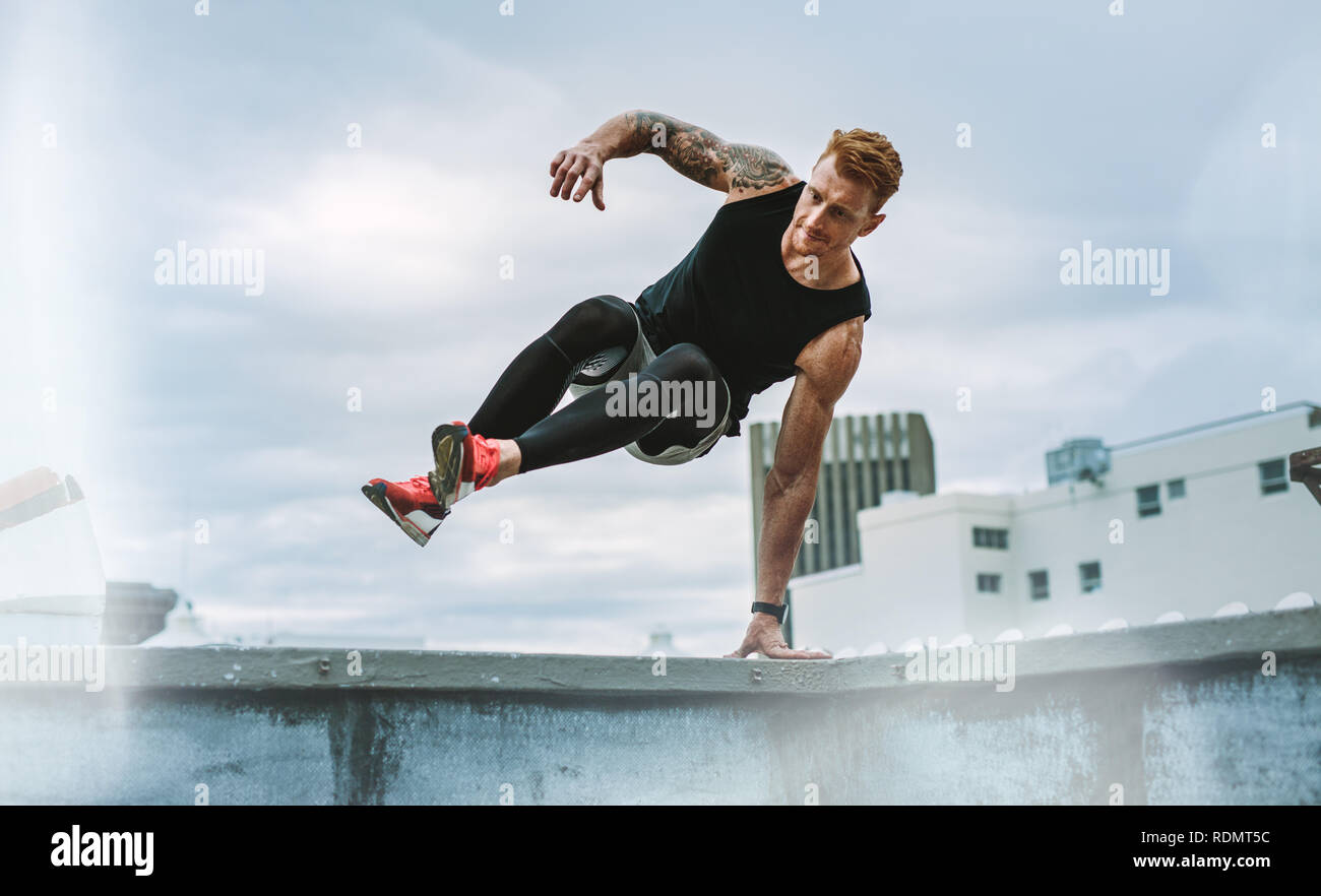 fitness man jumping on to the rooftop from the roof fence taking support of one hand. Man in fitness wear jumping from the rooftop fence on to the ter Stock Photo