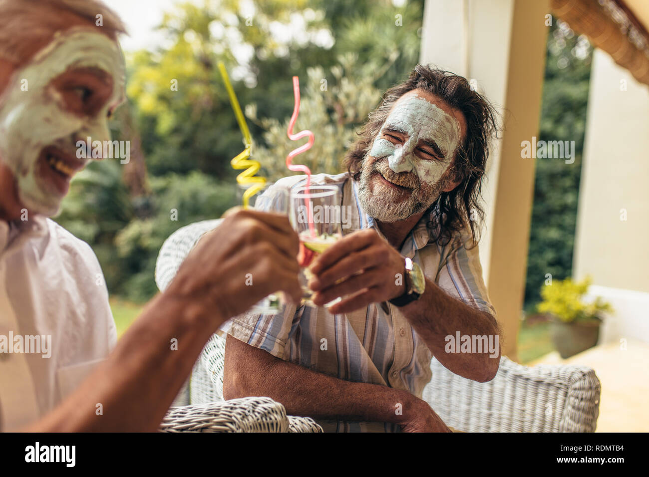 Happy old people toasting juice glasses with facial clay mask on. Funny senior men sitting together with face mask having juice. Stock Photo