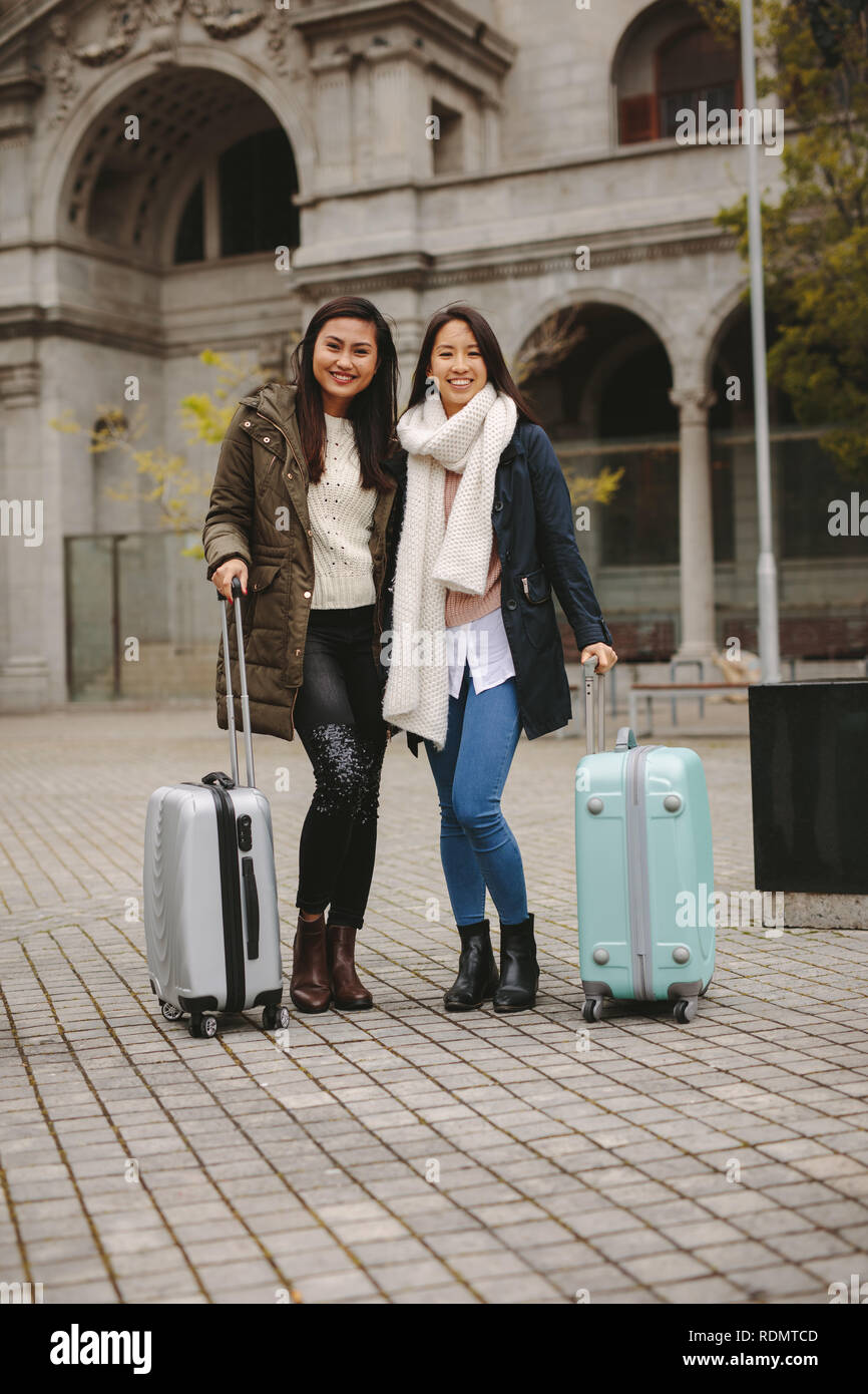 Tourist women standing in street holding their luggage bags. Smiling female tourists in winter clothes going around the city. Stock Photo