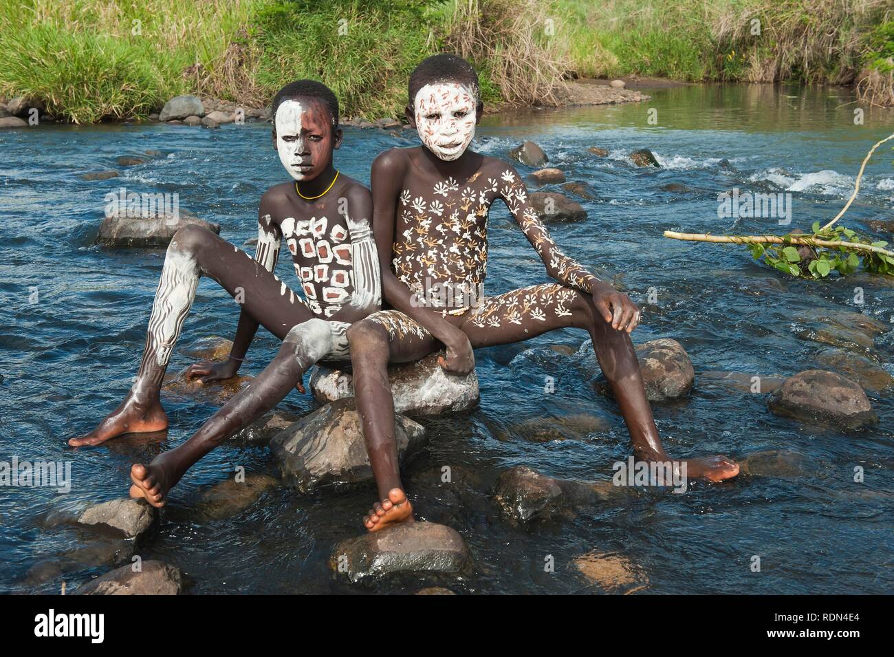 Two Surma boys with facial and body painting sitting on a rock in the river, Kibish, Omo River Valley, Ethiopia, Africa Stock Photo
