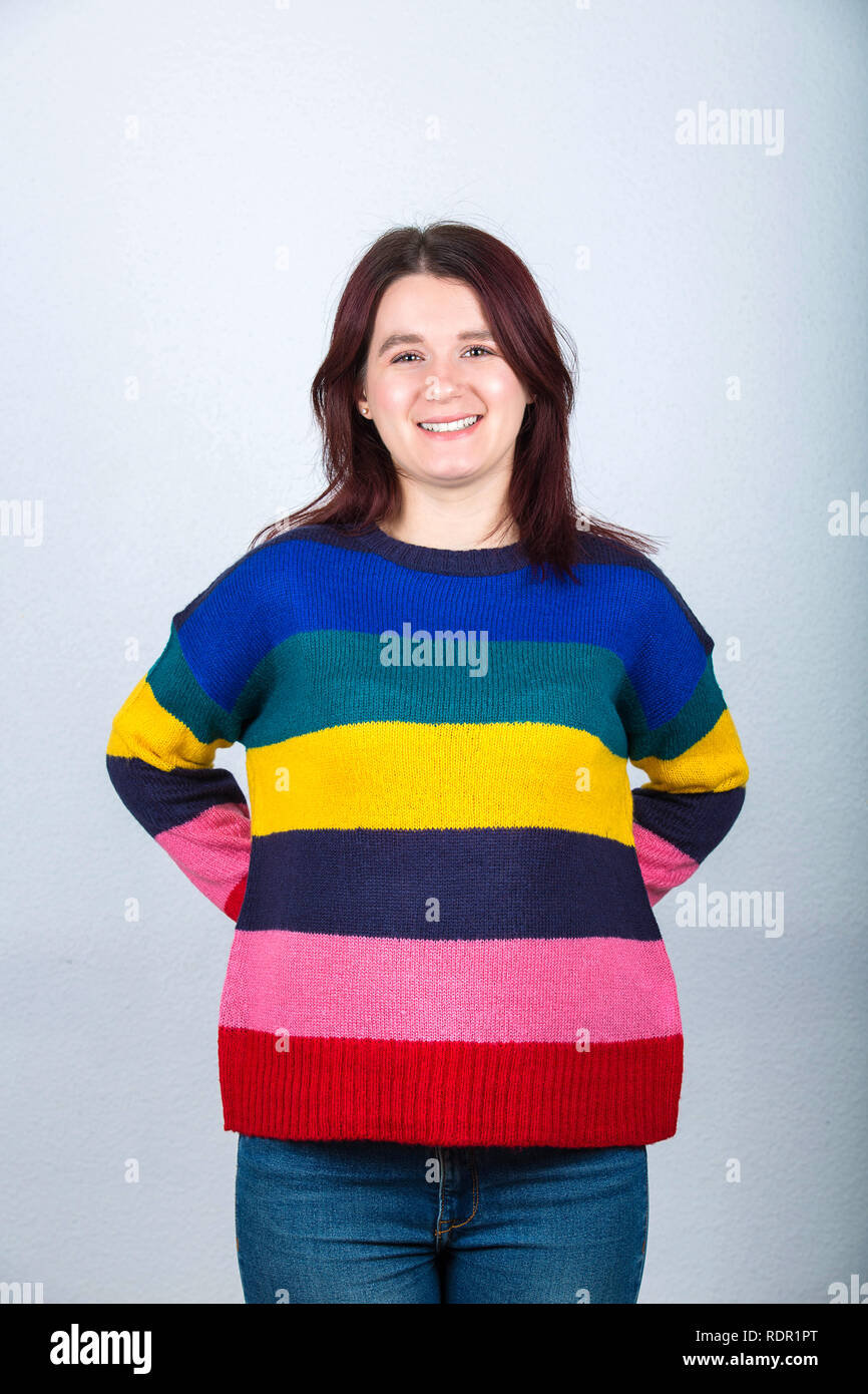 Standing young woman wearing jeans and colorful sweater, hands holding behind back, like hiding something or prepare a surprise isolated on grey backg Stock Photo