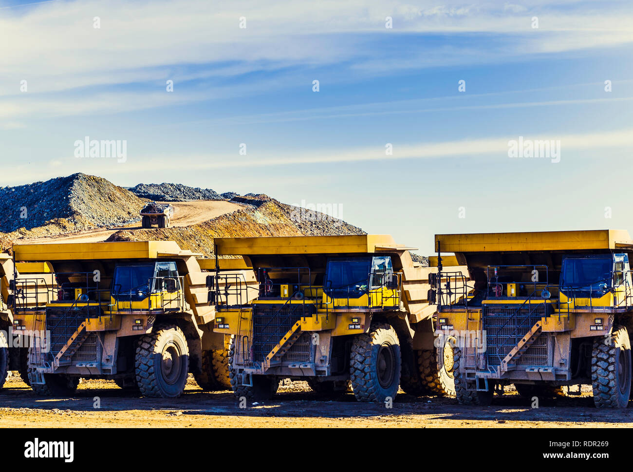 Big yellow dumper trucks placed in a row in the mine with dumper trucks laden with ore on the quarry road with blue sky and clouds in the background Stock Photo