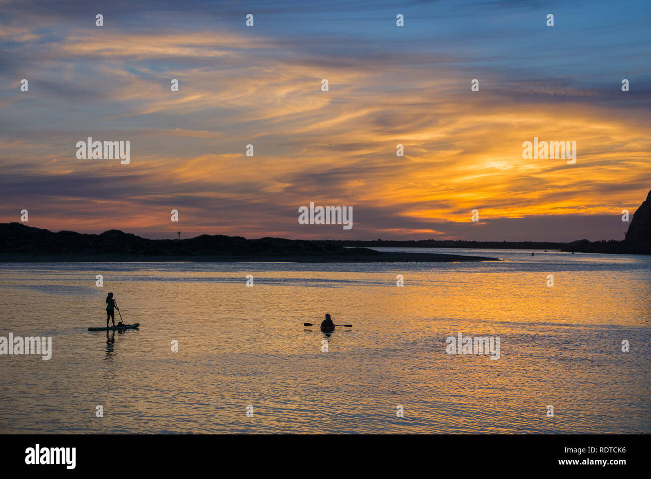 Colorful sunset as seen from the Morro Bay harbor; silhouettes of people enjoying the view from the kayak and paddle board; California Stock Photo