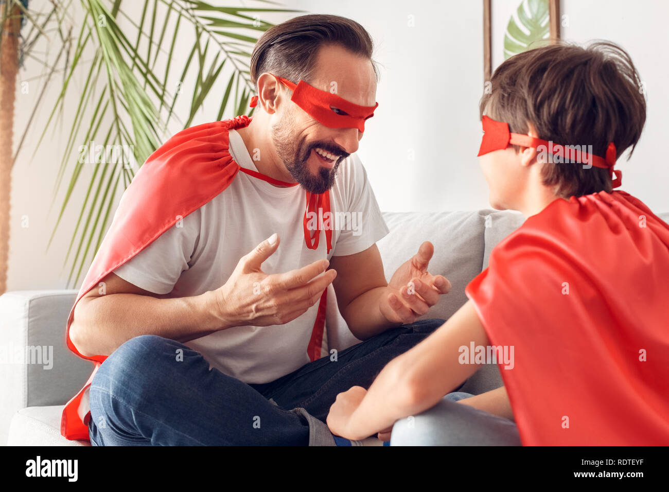 Father and son in superheroe costumes at home sitting on sofa talking laughing cheerful boy back close-up Stock Photo