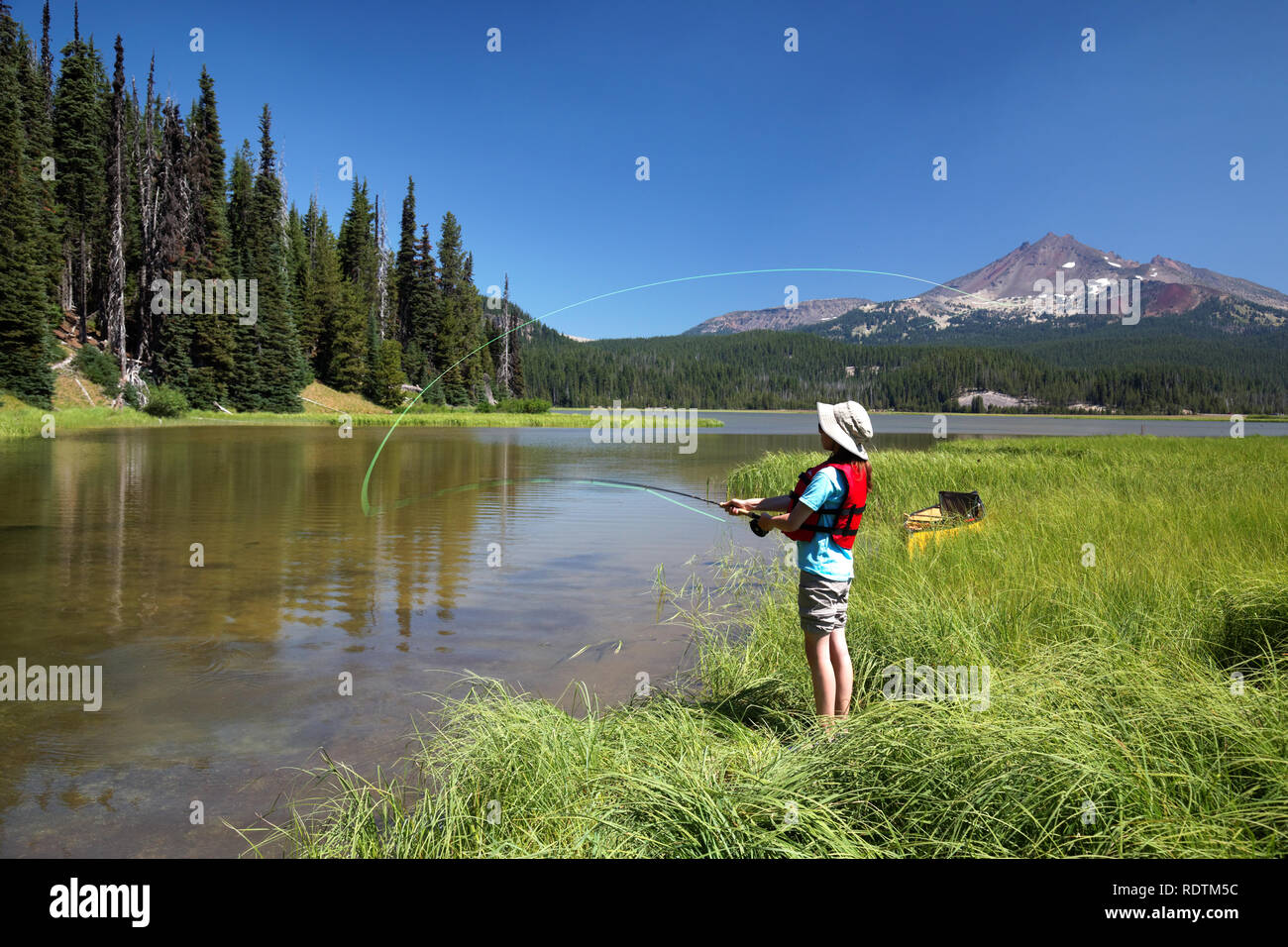 Girl fishing from shore along channel at Sparks Lake, Broken Top Mountain in background, Cascade Lakes, Oregon, USA Stock Photo