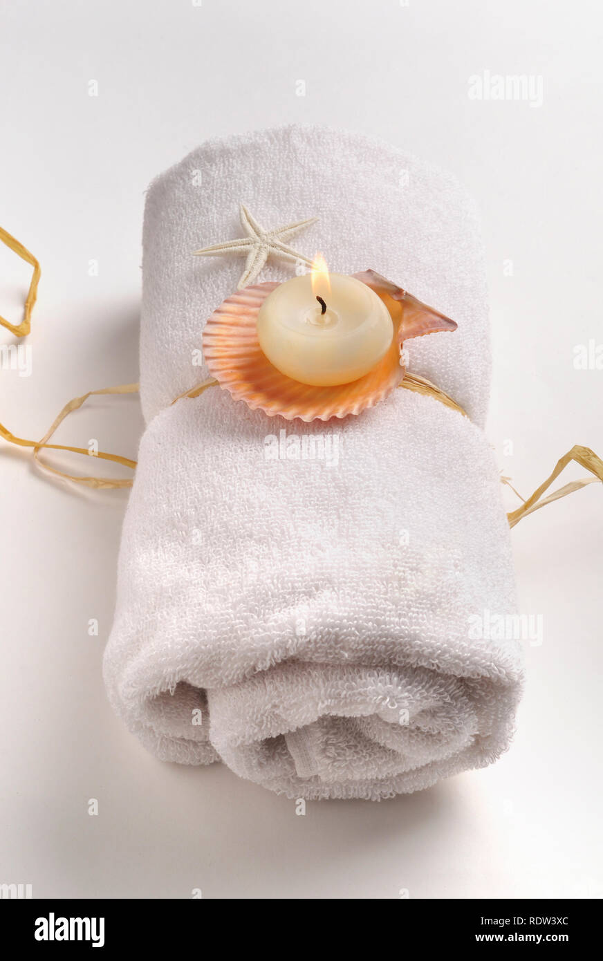 Lit-up scented candle with natural shells placed on rolled white towel - spa still life Stock Photo