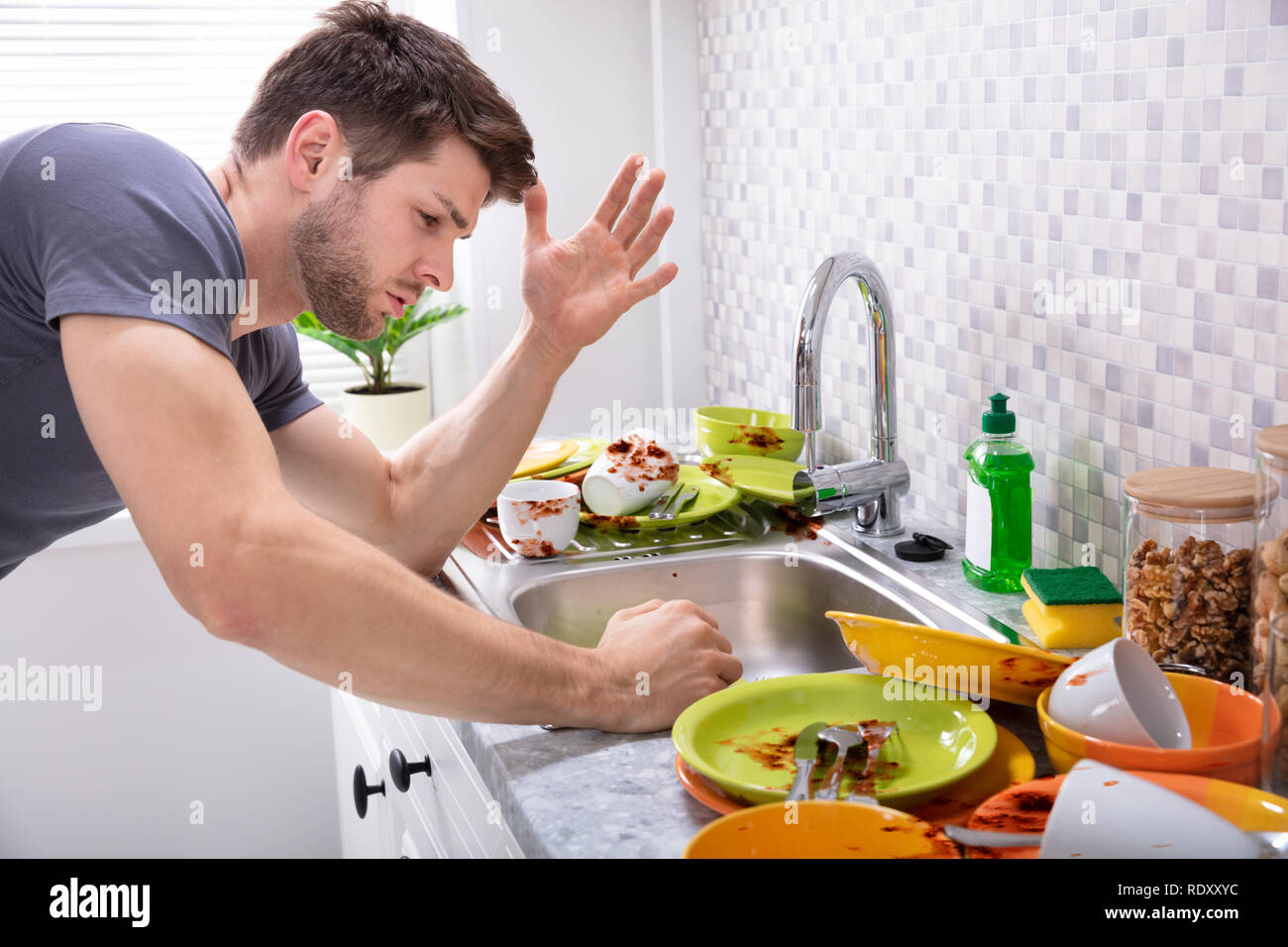 Sad Young Man Looking At Dirty Utensils Near Sink In Kitchen Stock Photo