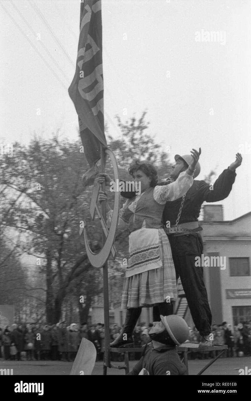 It was demonstration on 1-st May of 1978. Motorcyclist carries symbol of the Soviet era-the worker and the peasant. They stand upright on pedestal made directly on sidecar. The man and woman are specially dressed in appropriate clothes. They hold the flag of the USSR and metal hammer and sickle. The motorcyclist rides ahead of long column. The procession passes through the Central square of small town. Citizens with a sinking heart are look at the living symbol of communism. Stock Photo