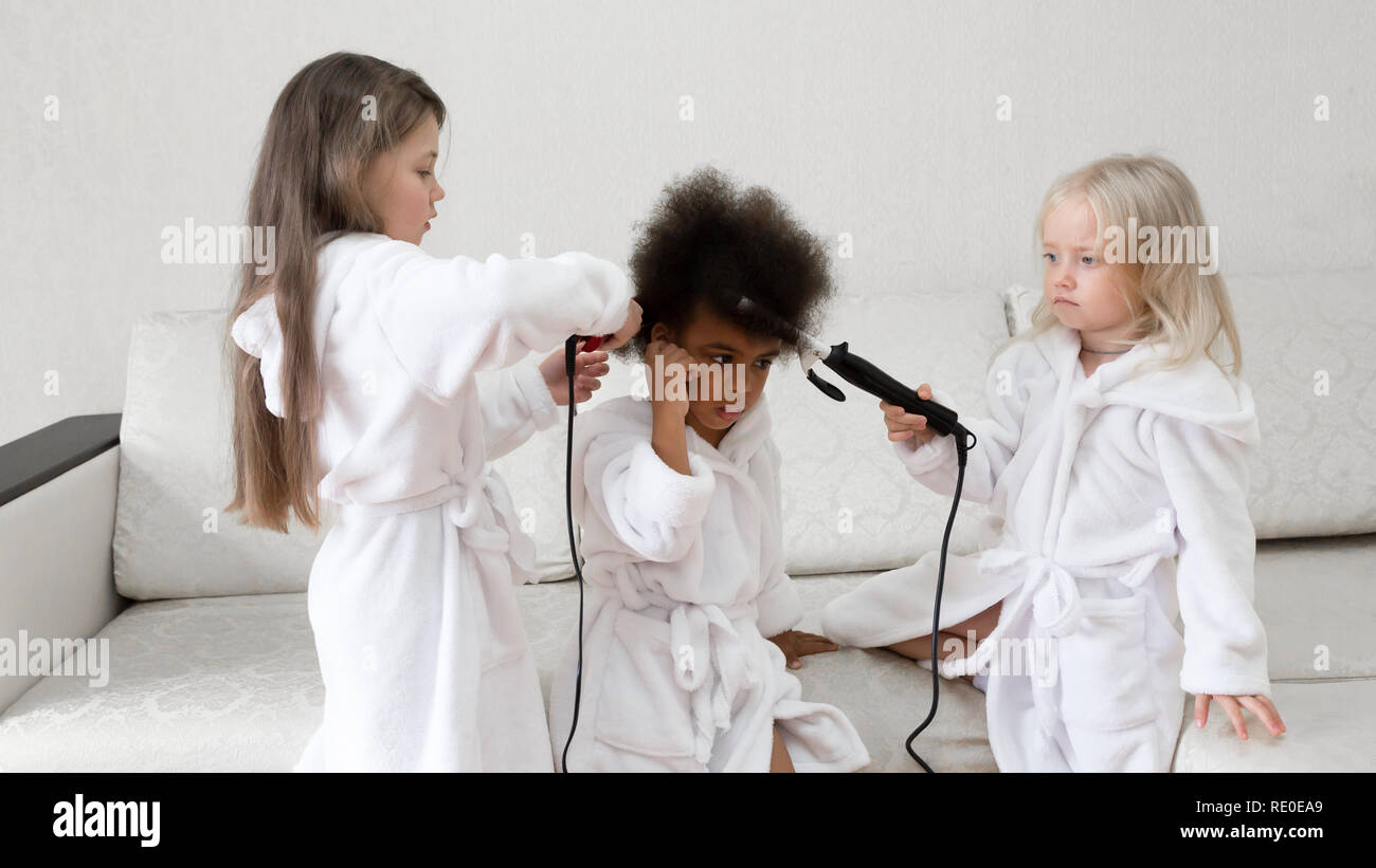 Children of different nationalities play together. Three girls friends in white coats twirl curls in each other's hair. Stock Photo