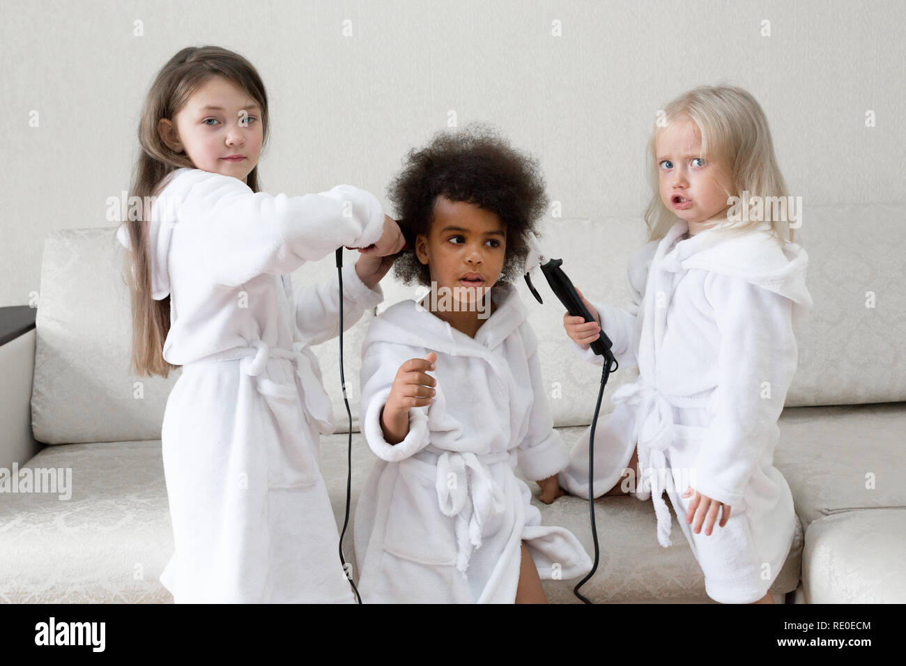 Children of different nationalities play together. Three girls friends in white coats twirl curls in each other's hair. Stock Photo