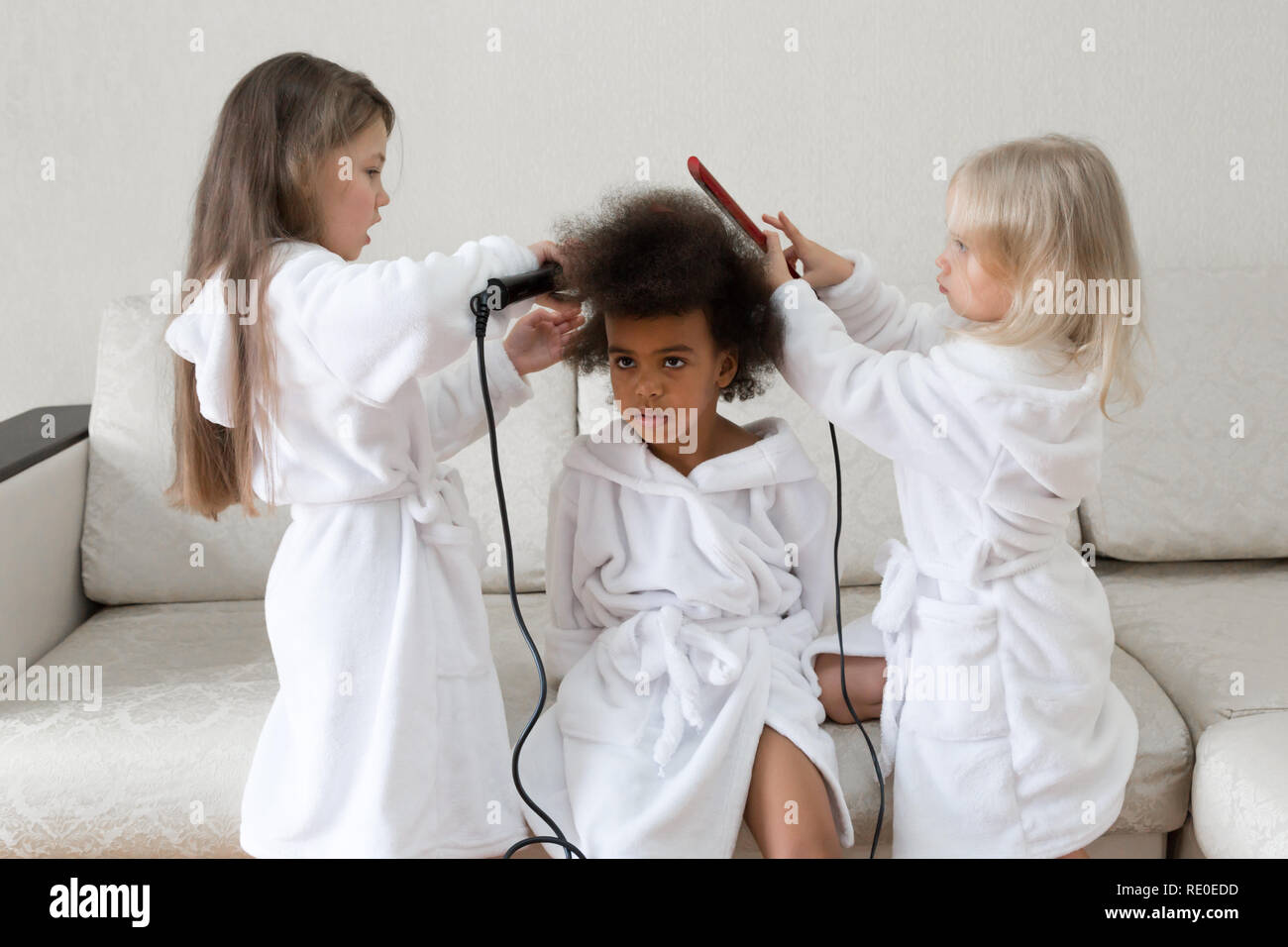Children of different nationalities play together. Three girls friends in white coats twirl curls in each other's hair. Stock Photo