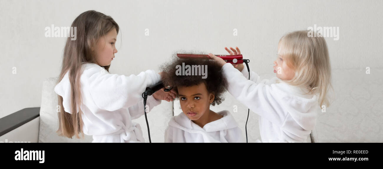 Children of different nationalities play together. Three girls friends in white coats twirl curls in each other's hair. Stock Photo