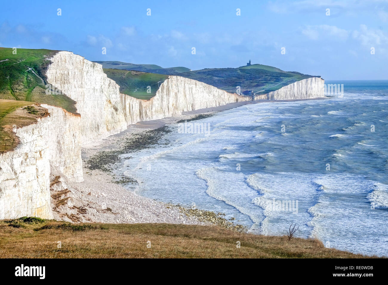 Seven Sisters cliffs, tall white chalk cliffs with the sea infront and on top of the cliffs is grass above a blue sky, at the bottom of the nearest cl Stock Photo