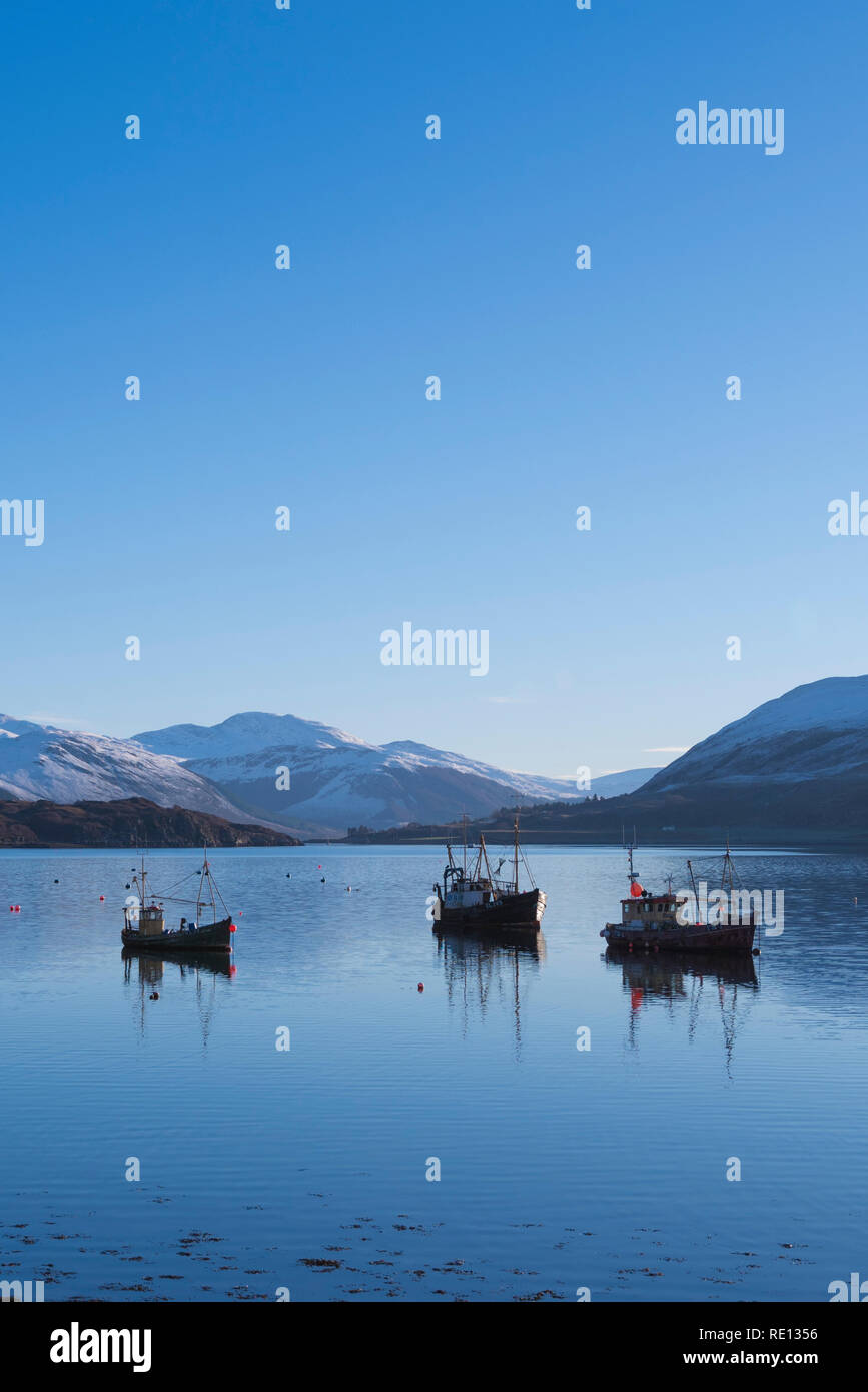 Fishing boats reflections moored in Loch Broom, Ullapool, with snowy winter mountains in the background Stock Photo