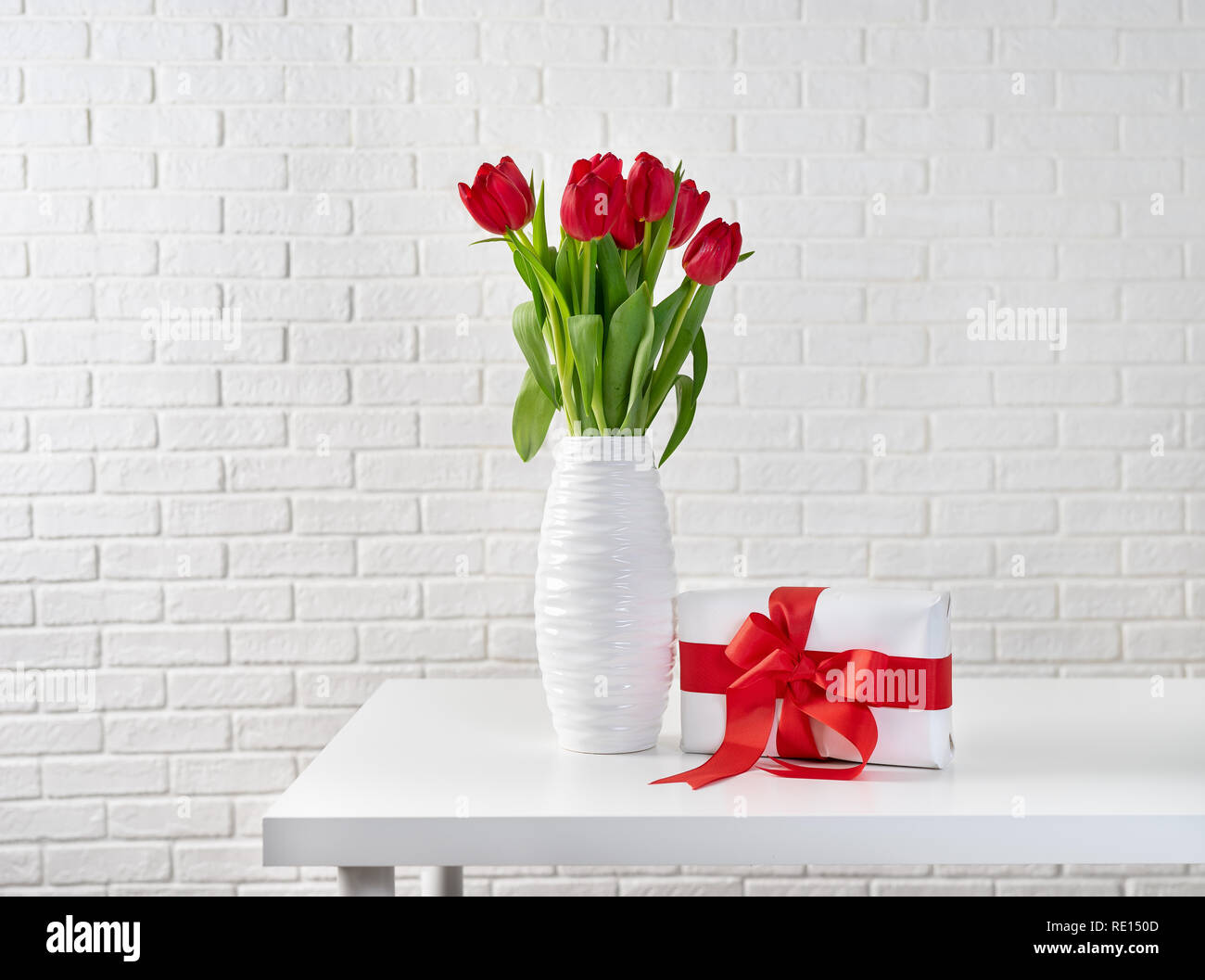 Red tulips in white vase next to a gift box with red ribbon Stock Photo