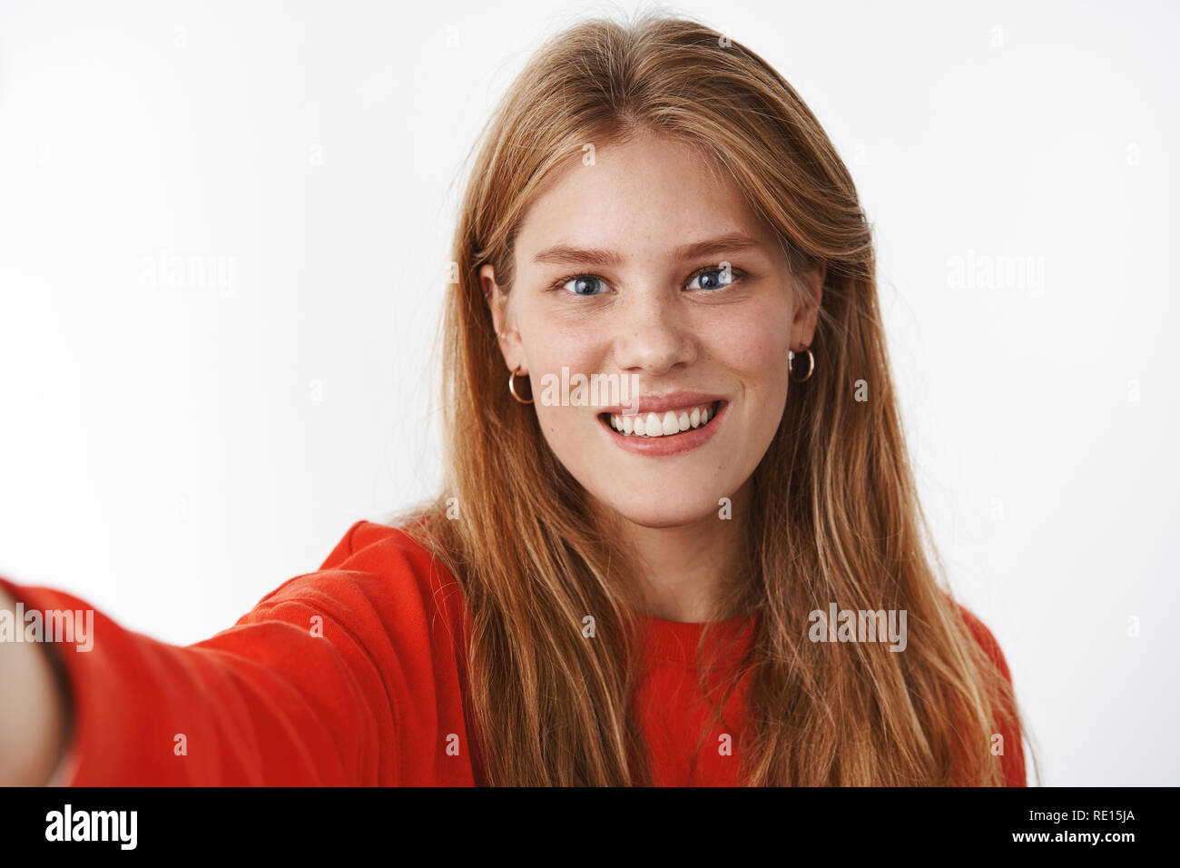 Close-up shot of nice tender young fair-haired girl with freckles, chubby cheeks and blue eyes pulling hand forward, holding camera and smiling as if  Stock Photo