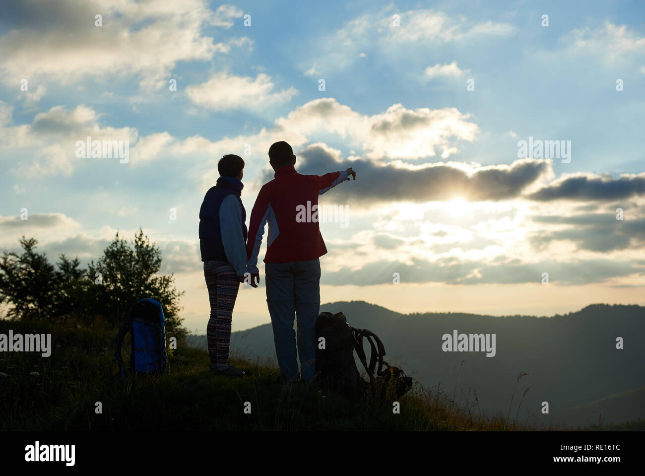 Silhouette of two tourists are standing on top of the mountain holding hands near backpacks against the backdrop of mountains and cloudy sky at sunset. Guy showing his hand in the distance. Rear view Stock Photo