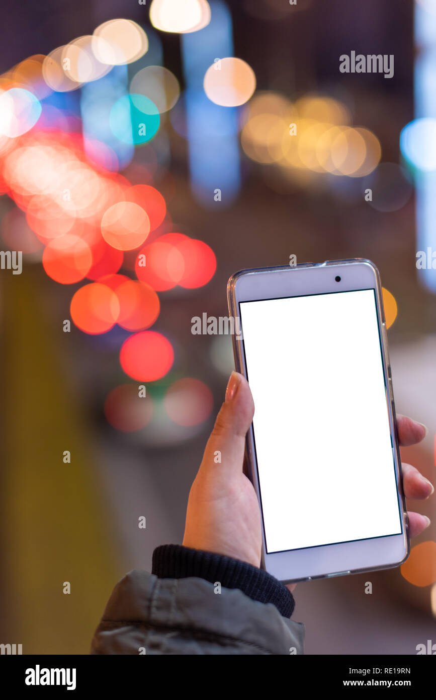 Woman holding a smartphone with a blank white screen at night with lights from car traffic in the background Stock Photo