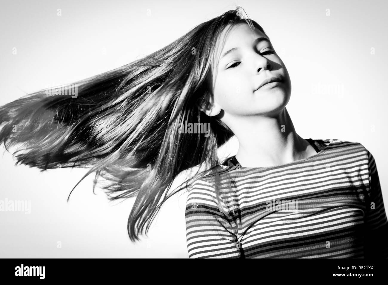 Tween girl is isolated against the sky while twirling and swinging her trendy pink tipped blond hair in black and white Stock Photo