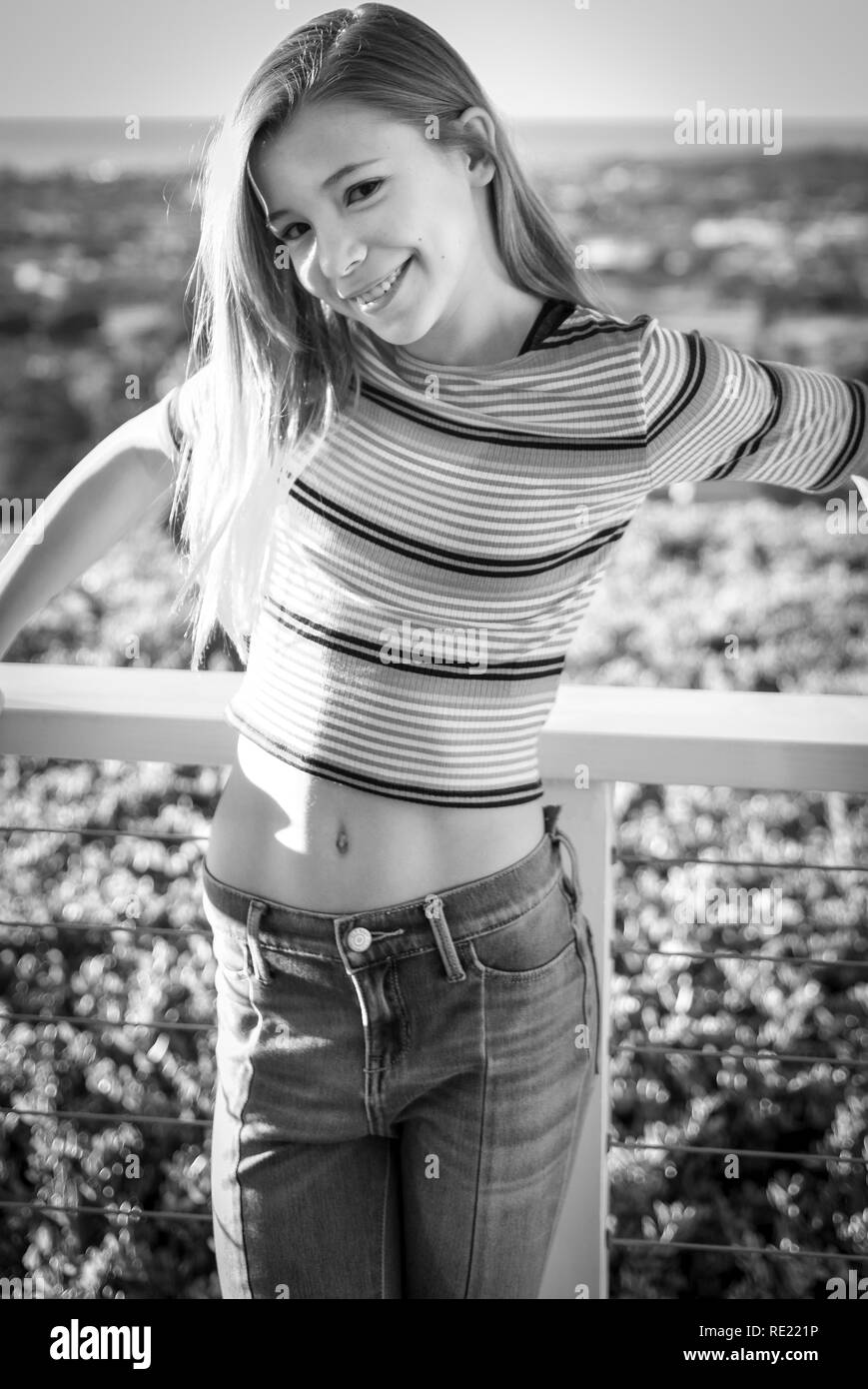 Cheeky tween girl gives knowing smile while leaning on railing of balcony with trendy pink hair and crop top showing abs in black and white Stock Photo