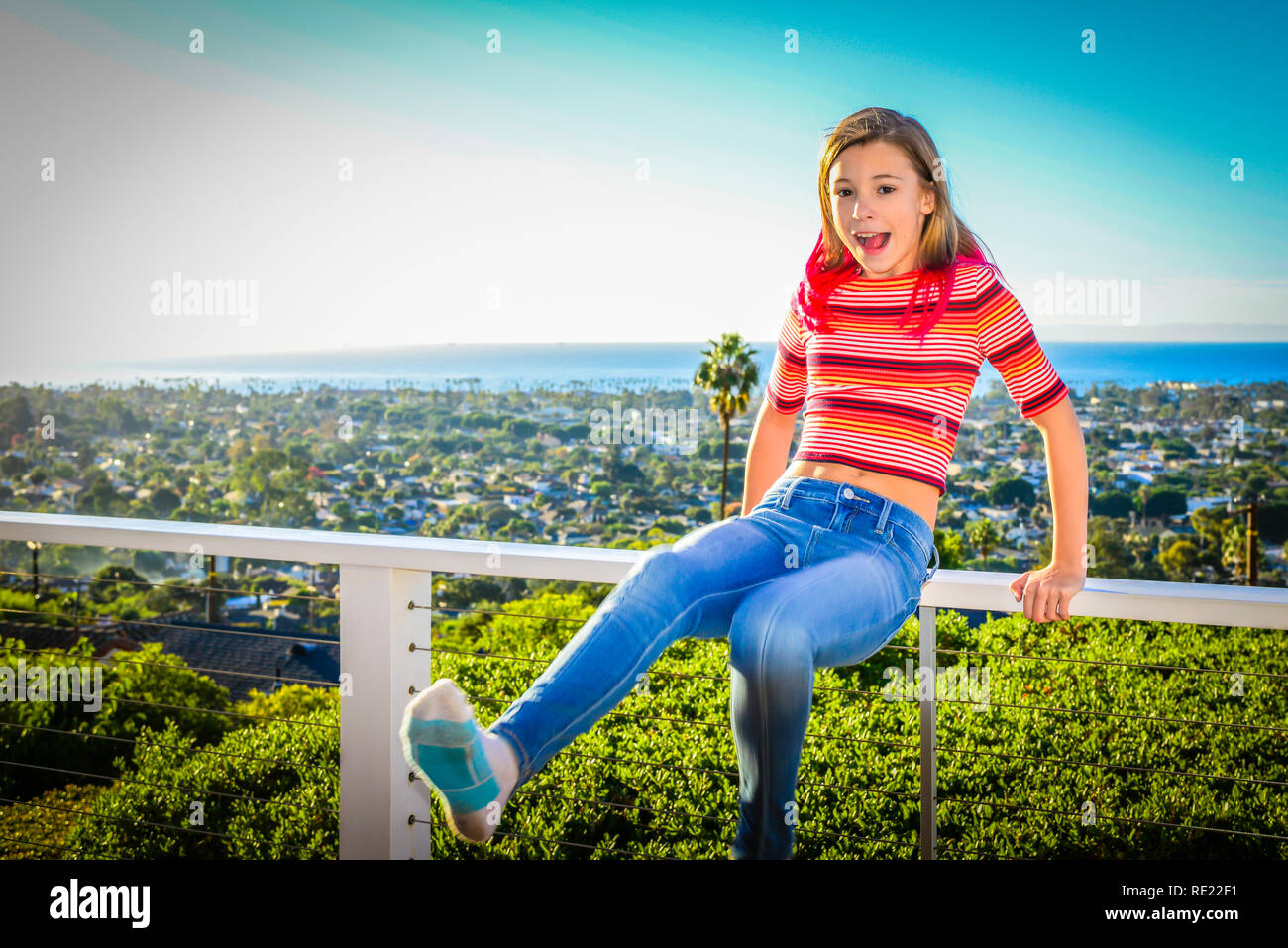 Young tween girl having dangerous fun balancing on railing of balcony overlooking a distant ocean Stock Photo