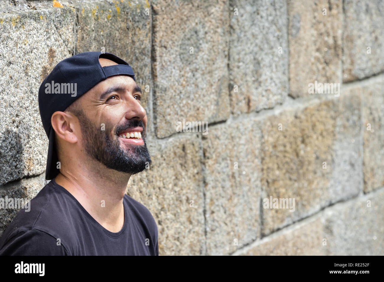 Portrait of a man with beard leaning on a stone wall, smiling, happy, dreaming, looking up away. Stock Photo