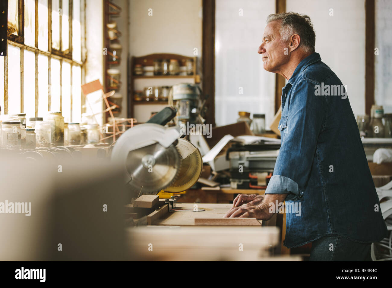 Side view of senior carpenter standing at the workbench and looking away. Thoughtful male carpenter in his workshop. Stock Photo