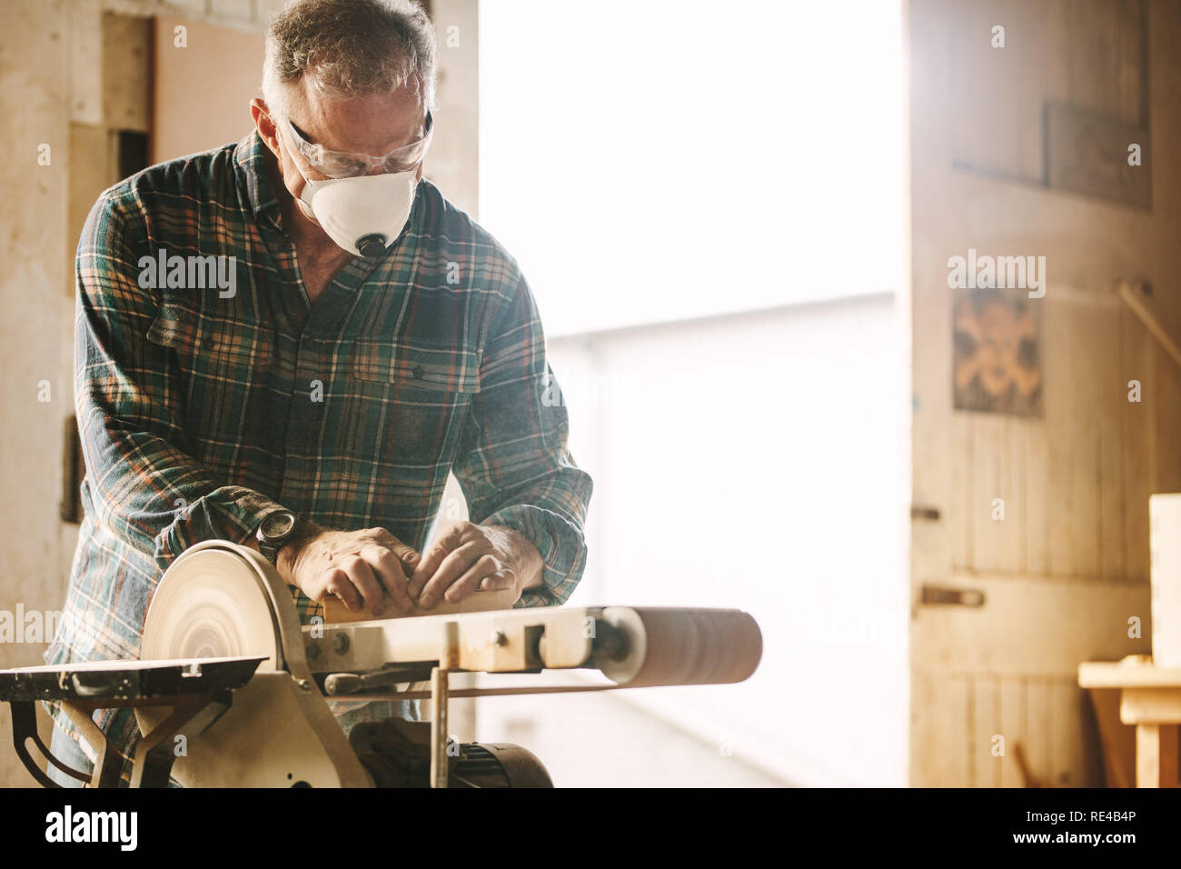 Senior carpenter with mask using belt sander. Carpenter sanding a wood with belt sander in carpentry workshop. Stock Photo