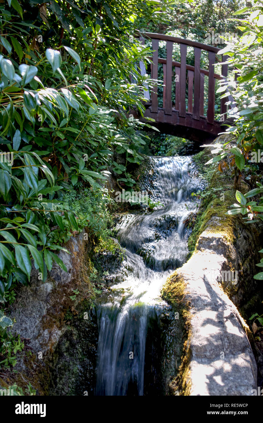 A small waterfall captured at a slow shutter speed with a small bridge in the background and a low wall to the side. The sun is shining on the waterfa Stock Photo