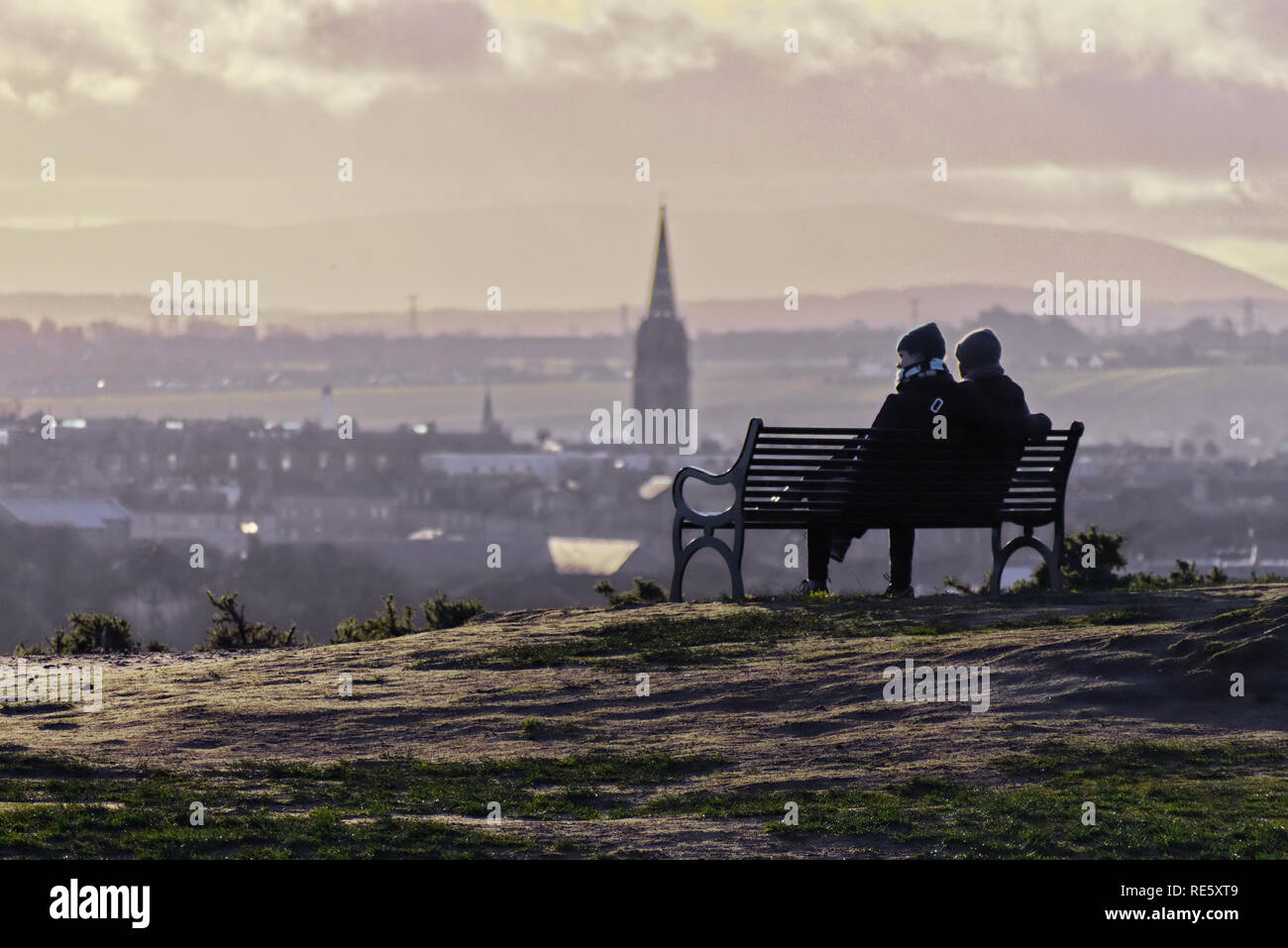 Two people sitting on a bench on top a hill at sunset in winter looking over the City of Edinburgh Stock Photo