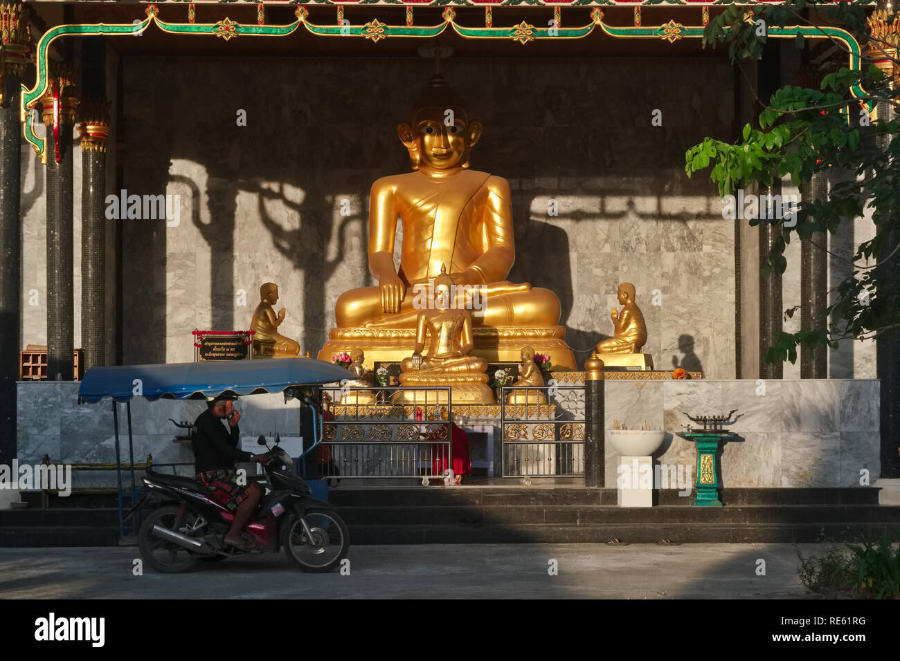 On an early morning,  man on a motorbike with sidecar passes a sun-lit golden Buddha statue at Wat Thepkassatri (Wat Don), Thalang, Phuket, Thailand Stock Photo