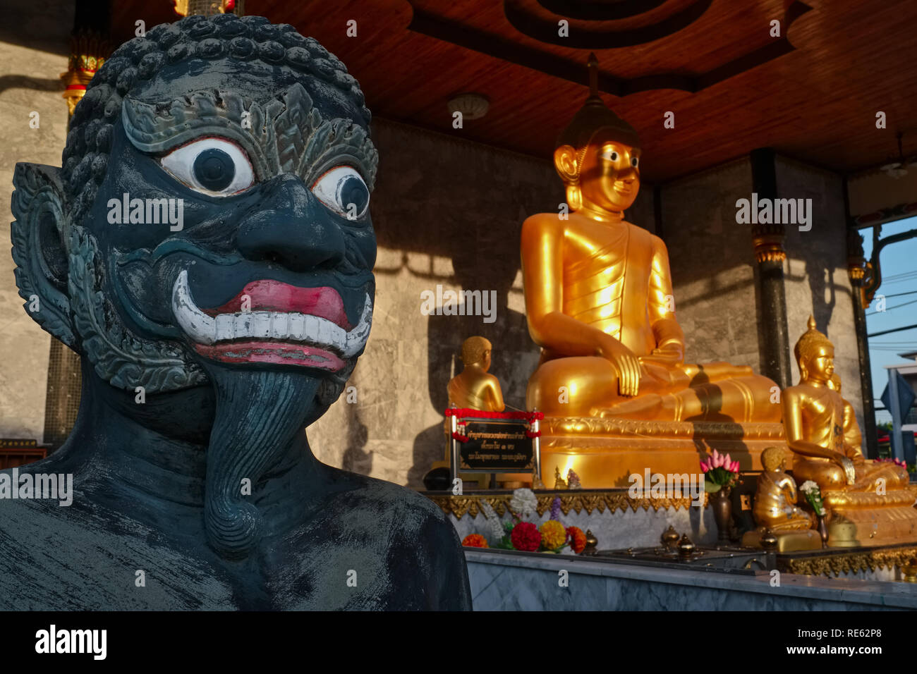 A demon statues stand next to a golden, sun-lit Buddha; on an early morning at Wat Thepkassatri (Wat Don), Thalang, Phuket, Thailand Stock Photo