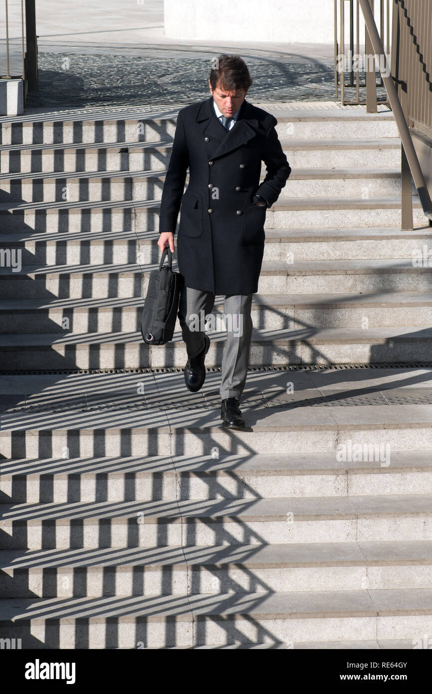 Businessman in coat walking down exterior concrete stairs with an interesting zigzag pattern from the shadow of the railing Stock Photo