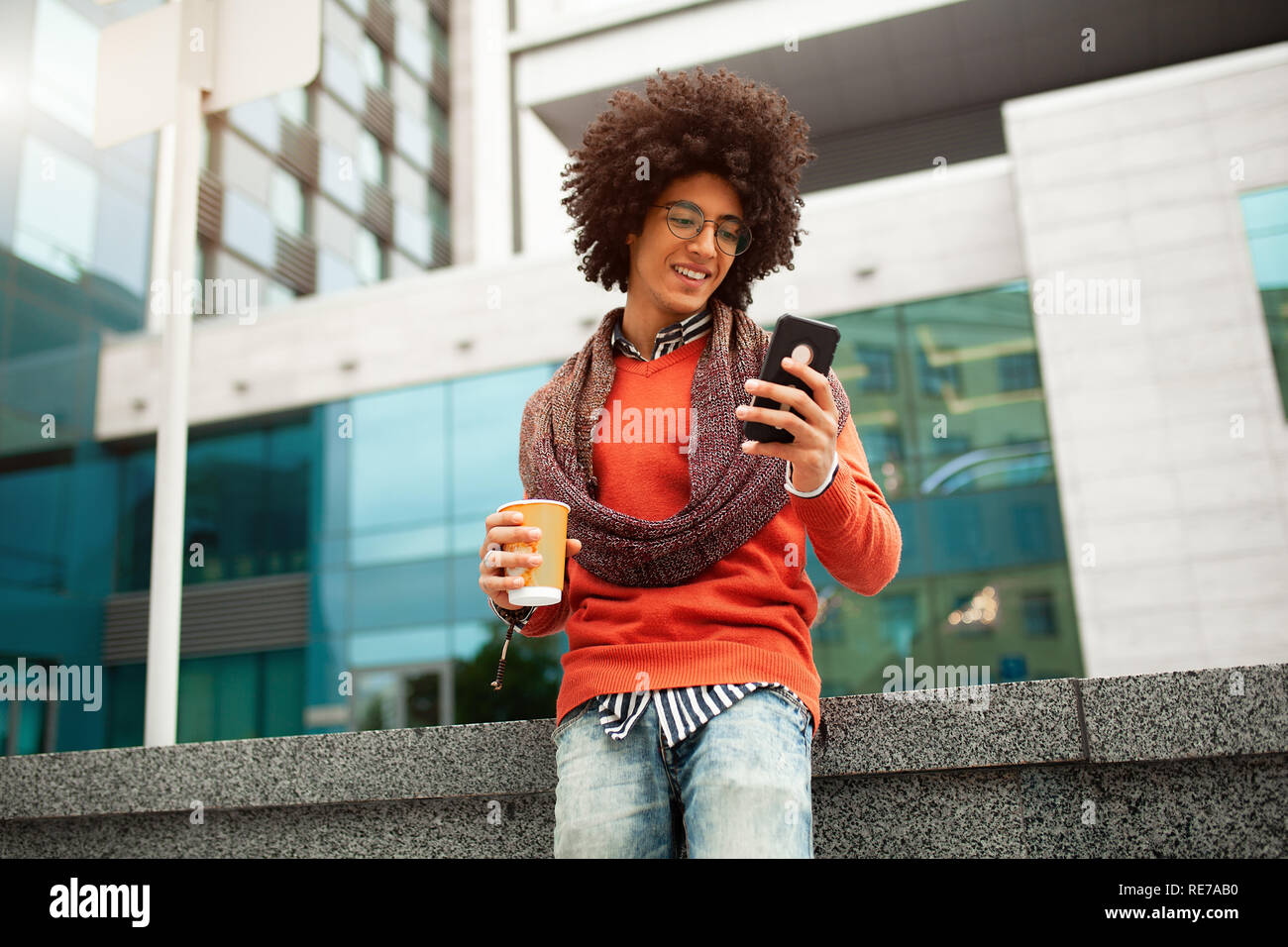 A handsome curly-haired guy of mixed race is dressed in a trendy youth-style scarf and sweater writes a message in a teleone against the background of Stock Photo