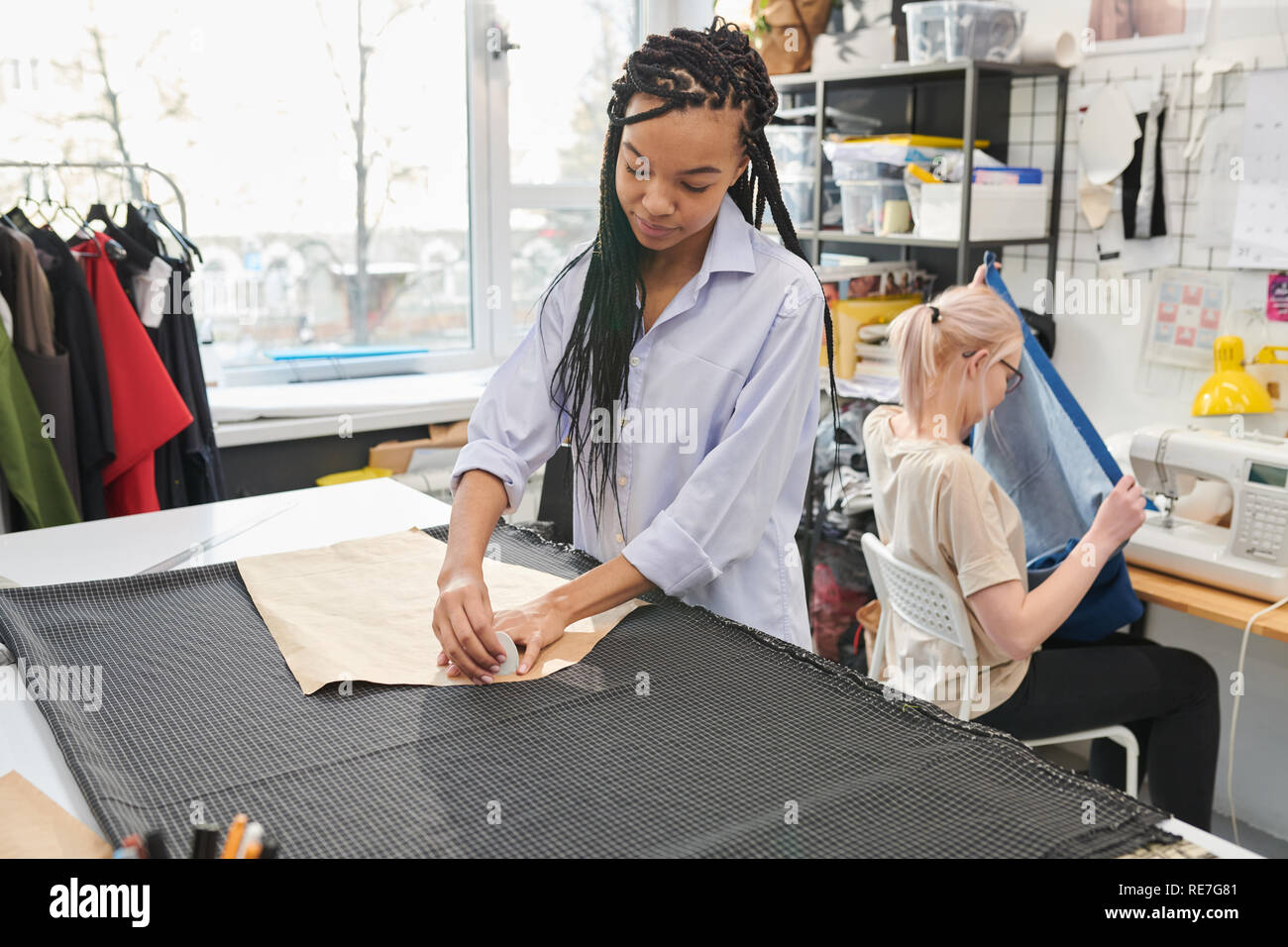 Young tailor working with fabric and pattern while her colleague using sewing machine on the background in workshop Stock Photo