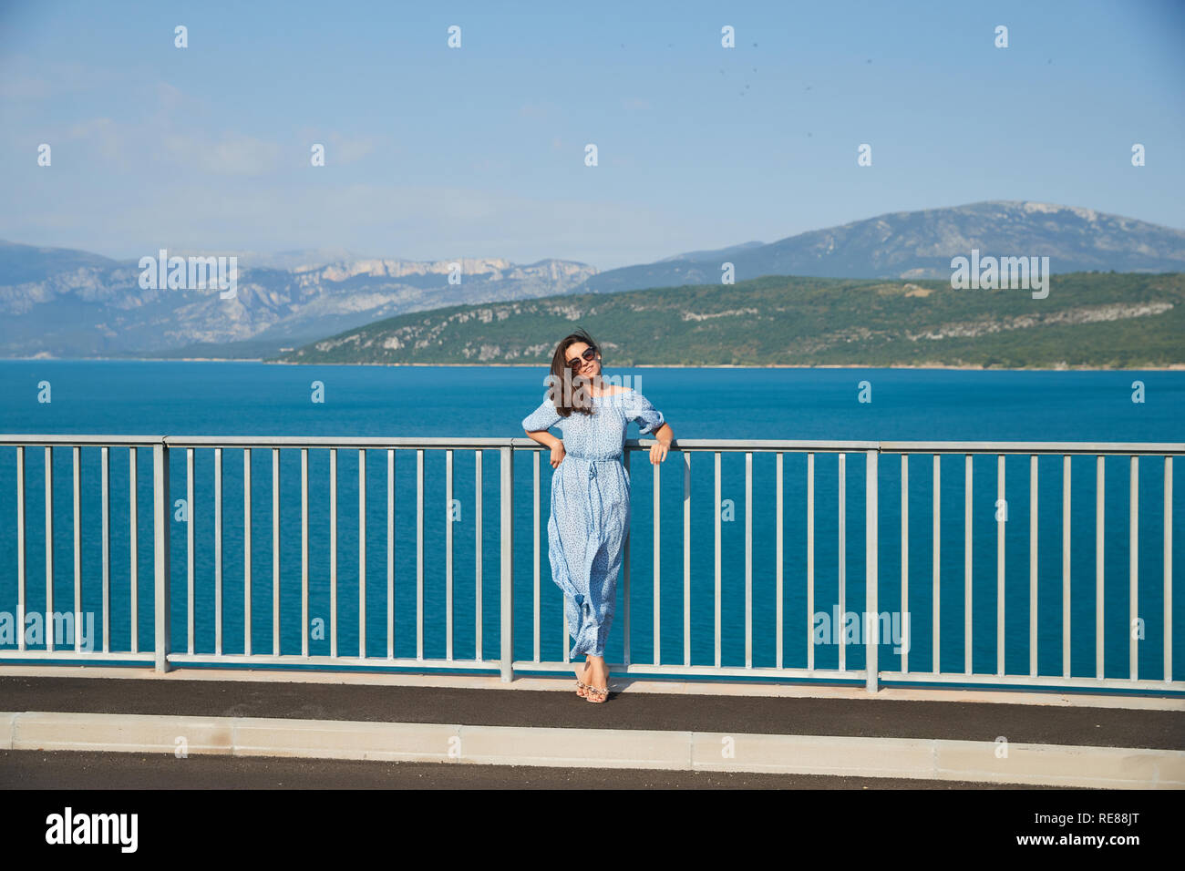 The beautiful girl in a blue dress and sunglasses poses on the bridge, the long chestnut hair, happy and smiles, azure water of the lake and slopes Stock Photo