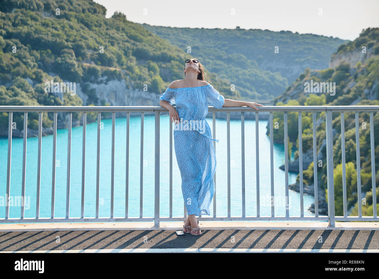 The beautiful girl in a blue dress and sunglasses poses on the bridge, the long chestnut hair, happy and smiles, azure water of the lake and slopes Stock Photo