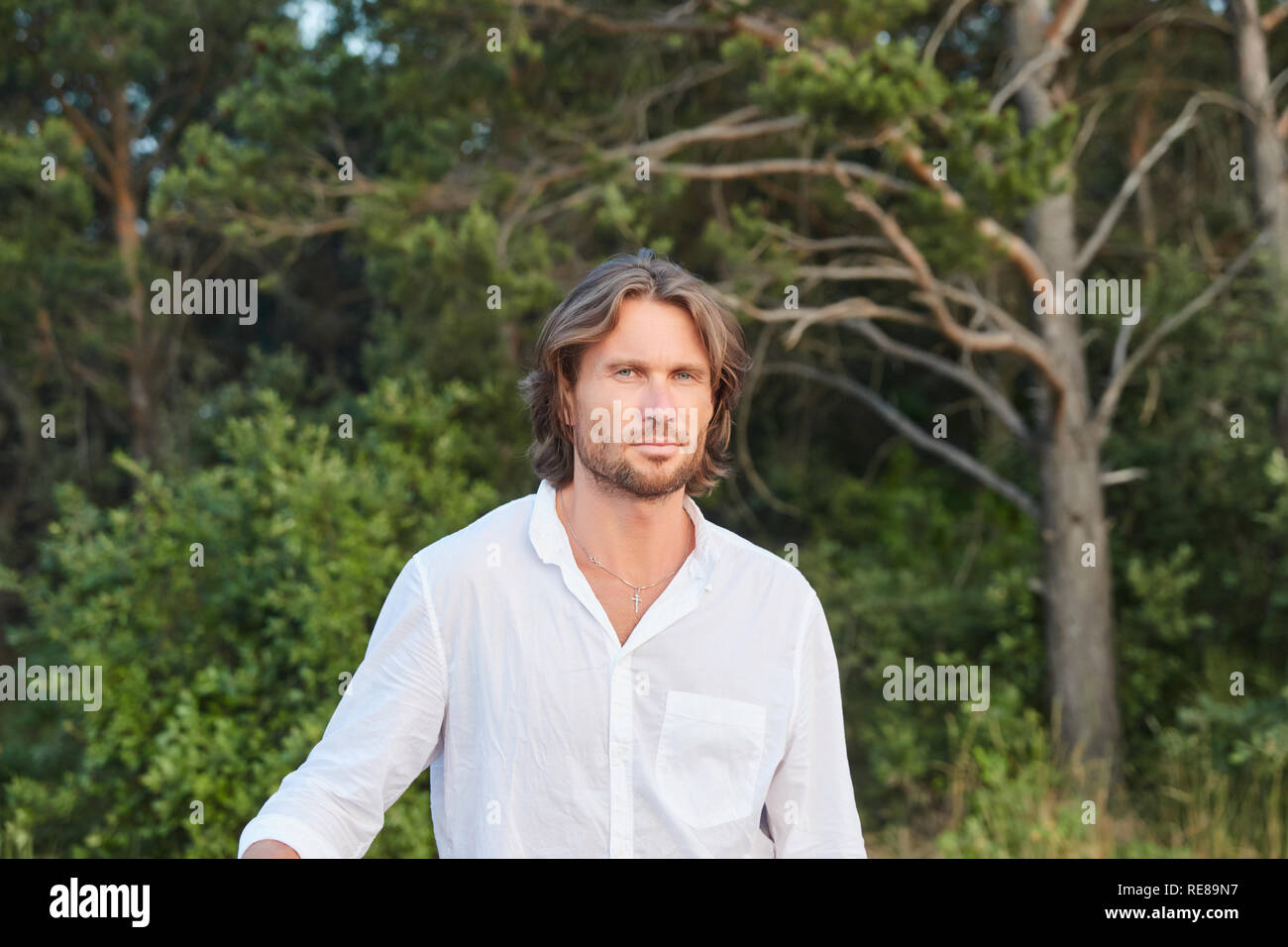 Portrait of the young handsome brutal man with long hair, the blue eyes, an easy bristle, he is dressed in a white shirt with a short sleeve Stock Photo