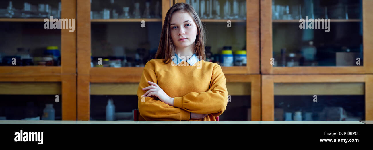 Portrait of young female college student in chemistry class, sitting behind the desk with crossed arms. Authentic Education concept banner. Stock Photo