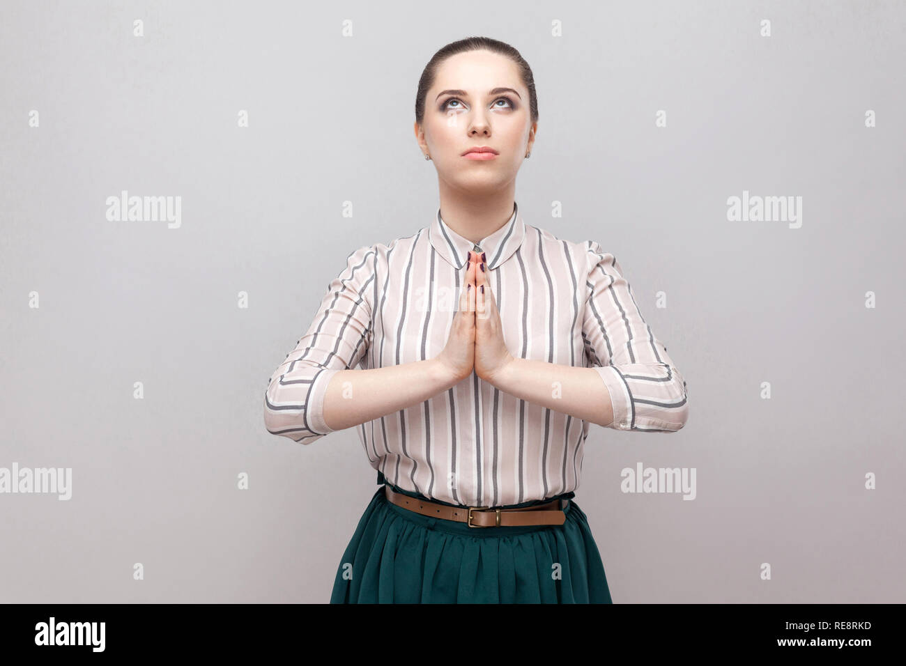 Portrait of hopeful beautiful young woman in striped shirt and green skirt with makeup and collected ban hairstyle, standing, looking uo and praying.  Stock Photo