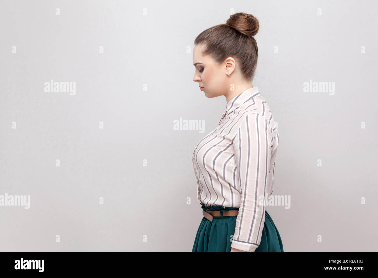 Profile side view portrait of unhappy beautiful young woman in striped shirt and green skirt and collected ban hairstyle, standing and holding her hea Stock Photo