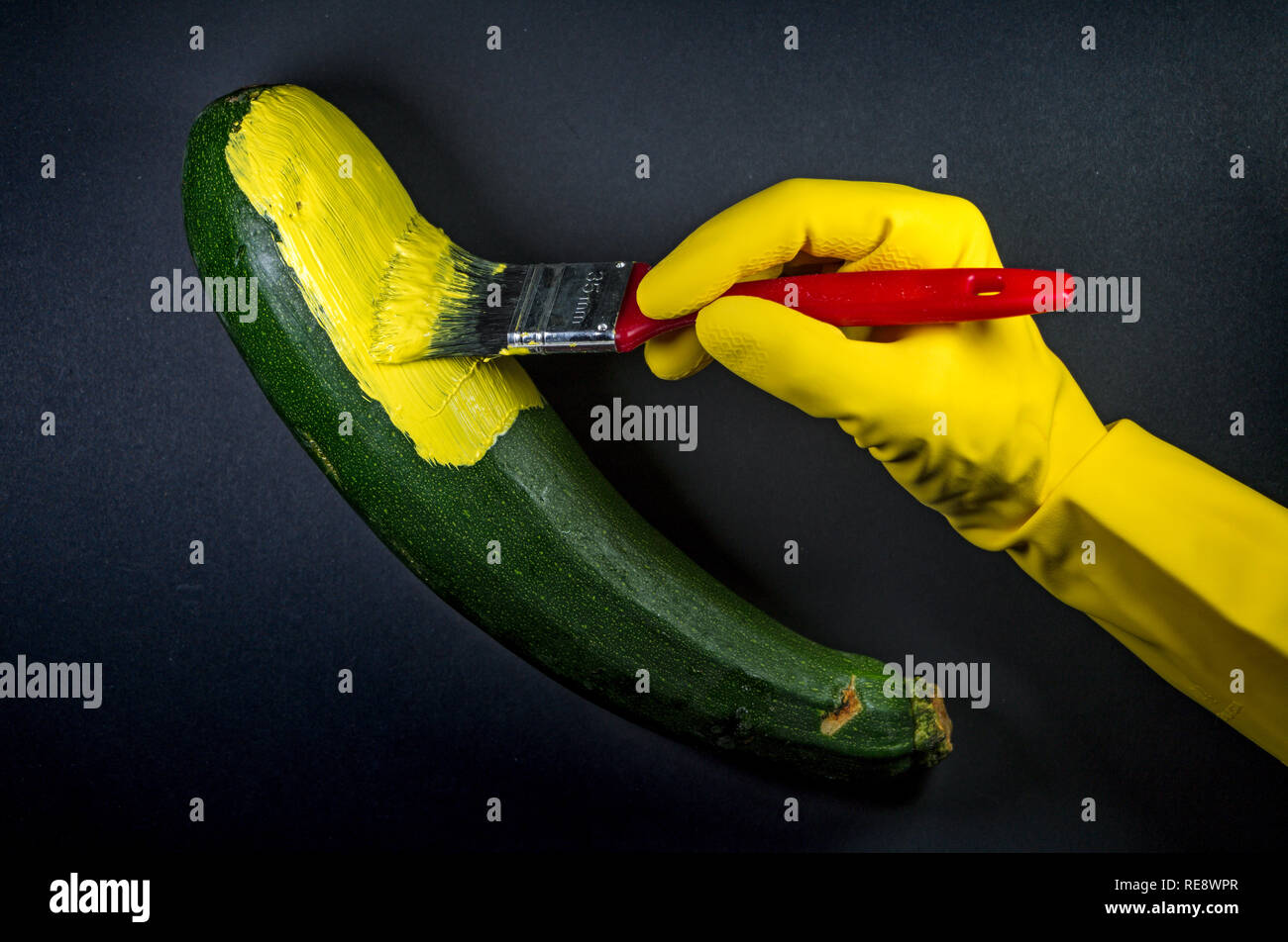 human hand in rubber gloves painting a zucchini yellow to look like a banana Stock Photo