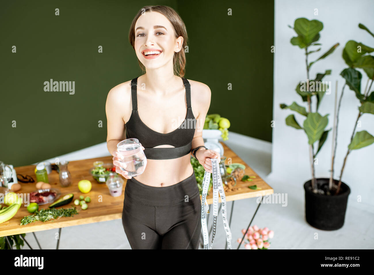 Happy woman in sportswear holding glass of water near the table full of healthy products. Healthy eating and sports lifestyle concept Stock Photo