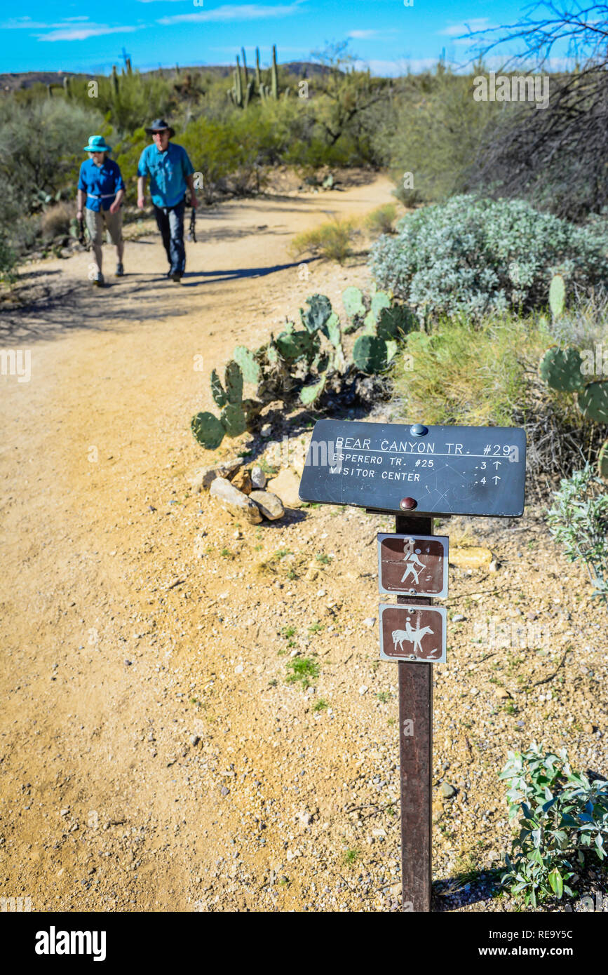 An older couple walks near a signpost with directions and distance for desert hiking trails in the Sabino Canyon Recreational Area in Tucson, AZ, USA Stock Photo