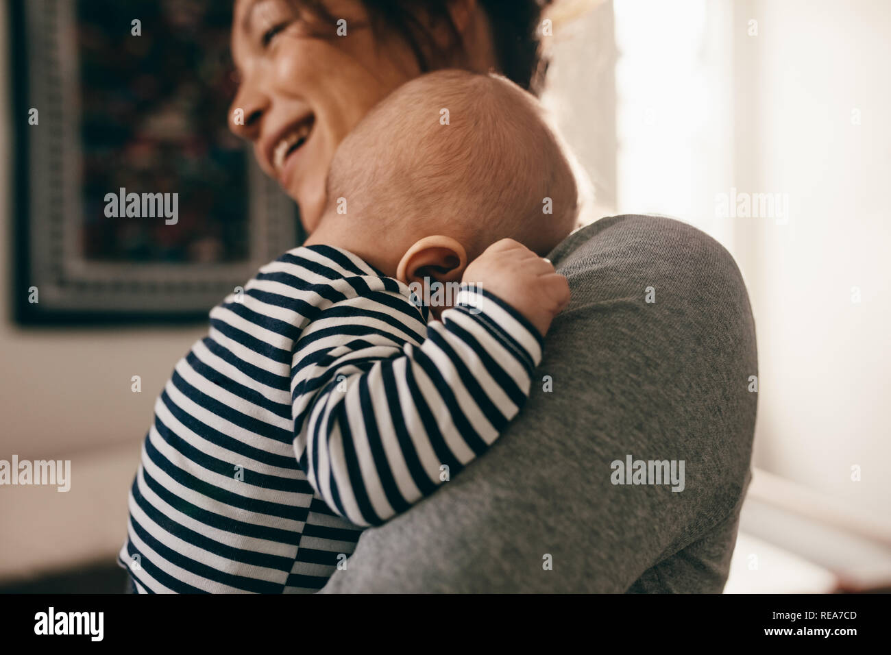 Side view of a smiling woman carrying her sleeping baby. Baby sleeping with his head resting on the shoulder of his mother at home. Stock Photo