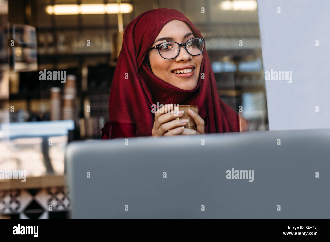 Pretty islamic girl in hijab and eyeglasses sitting at cafe with coffee in hand and looking away. Smiling muslim female at cafe with laptop. Stock Photo