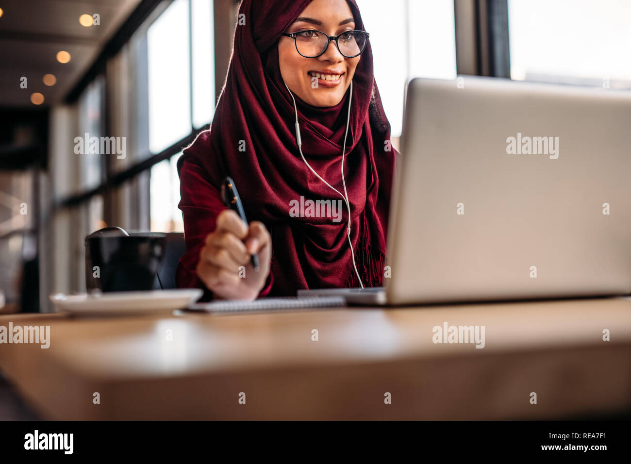 Woman in hijab sitting at cafe wearing earphones looking at laptop ...